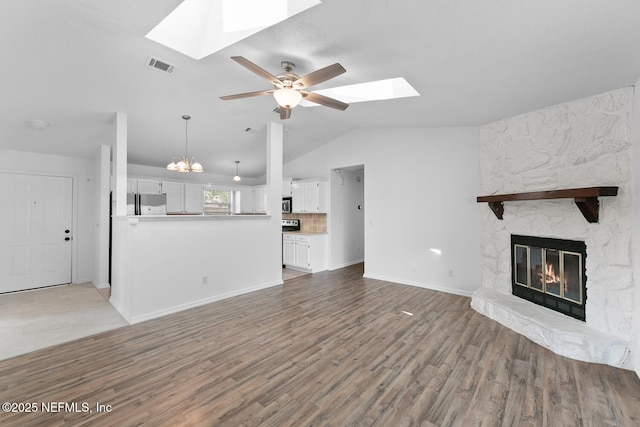 unfurnished living room featuring visible vents, a stone fireplace, wood finished floors, vaulted ceiling with skylight, and ceiling fan with notable chandelier