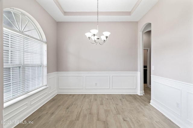 unfurnished dining area featuring arched walkways, an inviting chandelier, a raised ceiling, and light wood-style floors