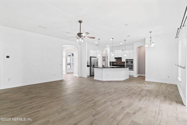 unfurnished living room with arched walkways, light wood finished floors, ceiling fan with notable chandelier, and baseboards