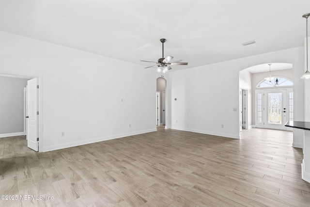 unfurnished living room with arched walkways, ceiling fan with notable chandelier, visible vents, baseboards, and light wood-type flooring