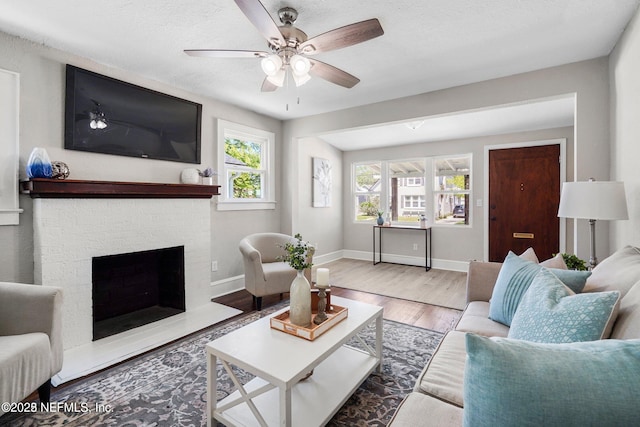 living room featuring a brick fireplace, ceiling fan, a textured ceiling, wood finished floors, and baseboards