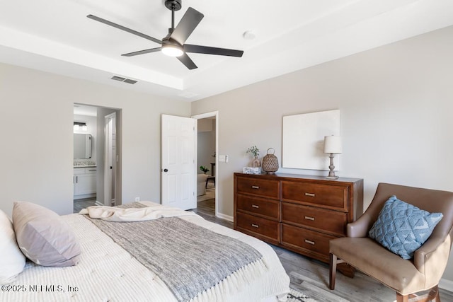 bedroom with a tray ceiling, visible vents, light wood-style flooring, connected bathroom, and ceiling fan