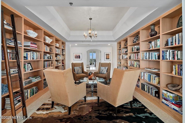 sitting room with wall of books, arched walkways, a raised ceiling, and a chandelier