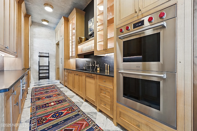 kitchen featuring stainless steel double oven, a sink, marble finish floor, dark countertops, and glass insert cabinets