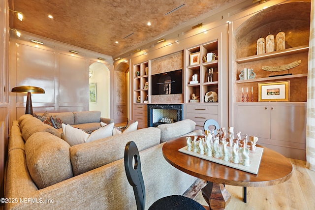 living room featuring arched walkways, wood finished floors, built in shelves, a decorative wall, and a fireplace