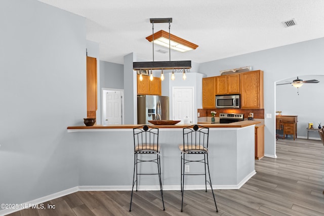 kitchen with light wood finished floors, a breakfast bar area, brown cabinets, decorative light fixtures, and stainless steel appliances