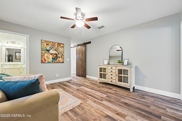 living room with ceiling fan, a barn door, wood finished floors, visible vents, and baseboards