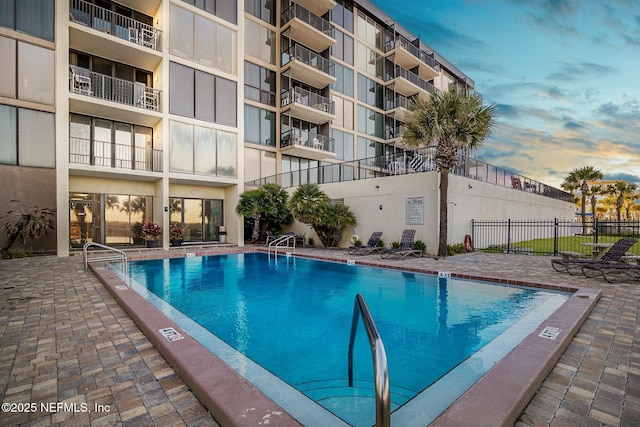 pool at dusk featuring a patio area, fence, and a community pool