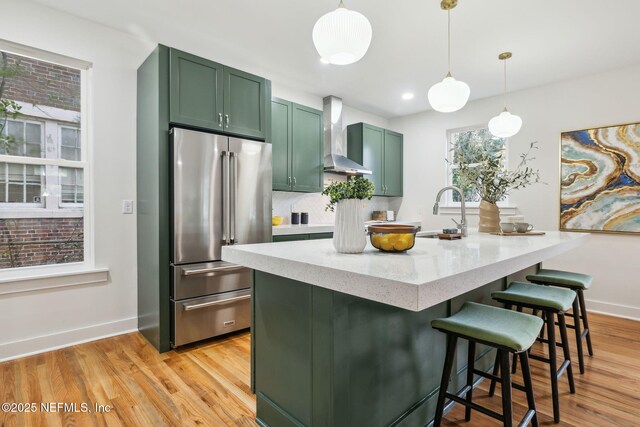 kitchen with high end fridge, a sink, wall chimney exhaust hood, light wood finished floors, and green cabinetry