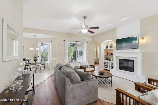 living area featuring arched walkways, a glass covered fireplace, ceiling fan, wood finished floors, and built in shelves