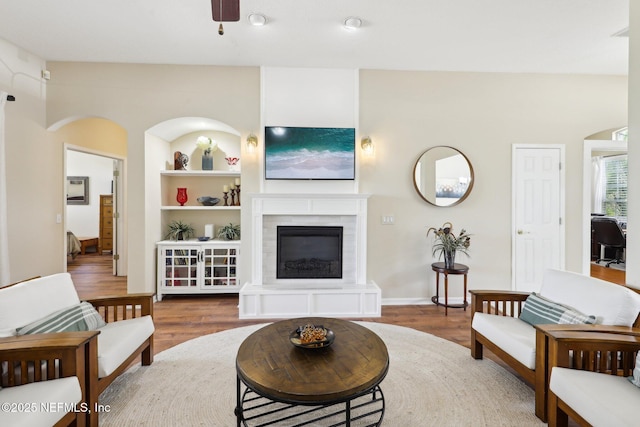 living room featuring built in shelves, a glass covered fireplace, ceiling fan, and wood finished floors