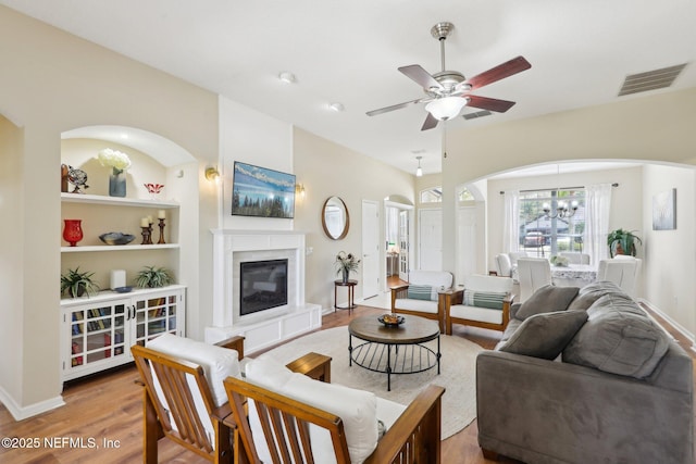 living room with visible vents, a glass covered fireplace, wood finished floors, baseboards, and ceiling fan with notable chandelier