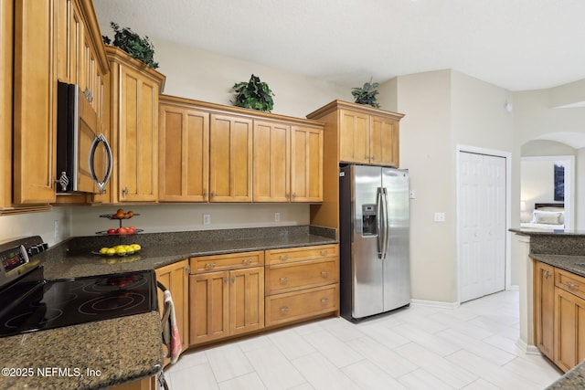 kitchen featuring arched walkways, stainless steel appliances, dark stone countertops, and brown cabinets