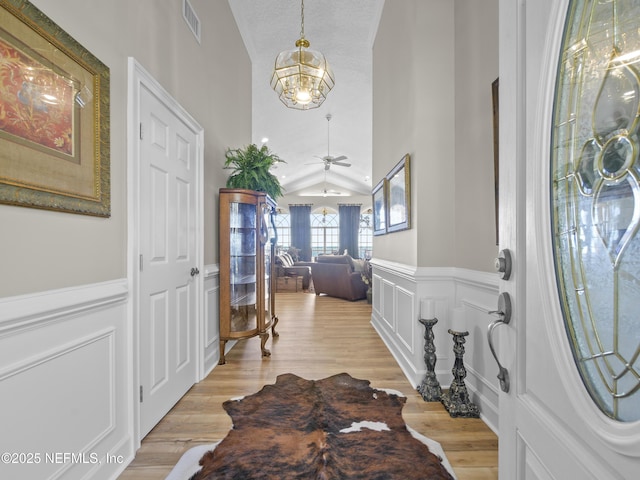 entryway featuring light wood-type flooring, visible vents, ceiling fan with notable chandelier, wainscoting, and vaulted ceiling