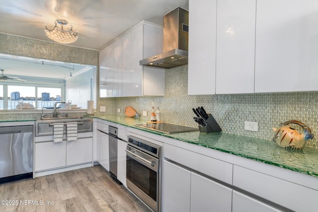 kitchen featuring light wood finished floors, white cabinets, appliances with stainless steel finishes, wall chimney range hood, and tasteful backsplash
