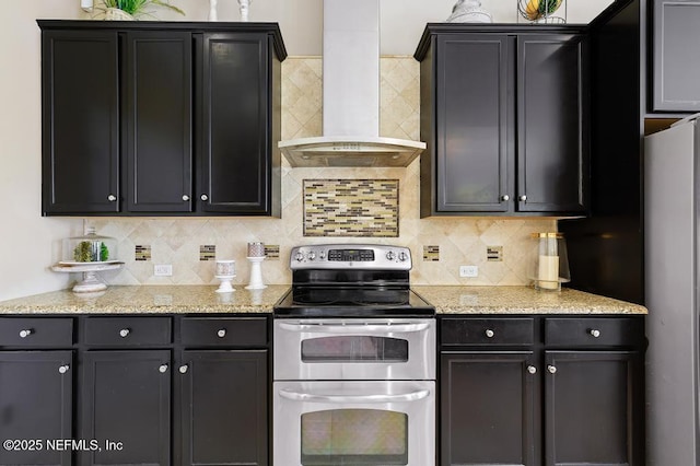 kitchen with light stone counters, dark cabinets, stainless steel appliances, backsplash, and wall chimney exhaust hood