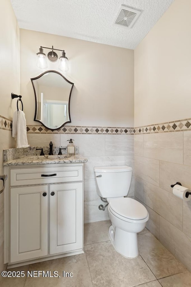 half bathroom with visible vents, toilet, a textured ceiling, vanity, and tile walls