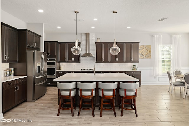 kitchen featuring appliances with stainless steel finishes, an island with sink, light countertops, and wall chimney range hood