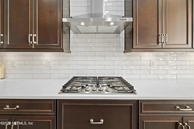 kitchen featuring stainless steel gas stovetop, wall chimney range hood, decorative backsplash, light stone countertops, and dark brown cabinets