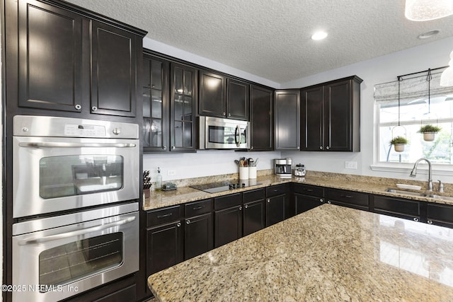 kitchen featuring a sink, a textured ceiling, stainless steel appliances, glass insert cabinets, and light stone countertops