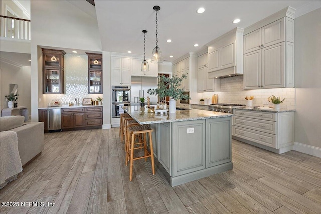 kitchen featuring an island with sink, light wood-style floors, appliances with stainless steel finishes, glass insert cabinets, and custom exhaust hood