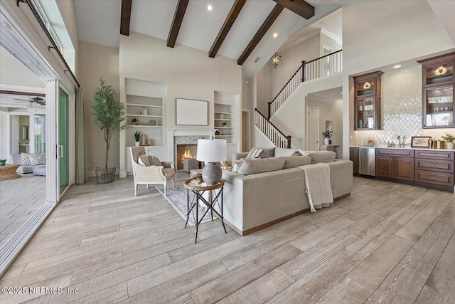 living room with beam ceiling, light wood-style flooring, a warm lit fireplace, wet bar, and high vaulted ceiling