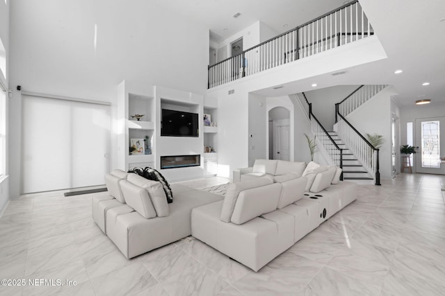 living area featuring built in features, arched walkways, a glass covered fireplace, marble finish floor, and stairs