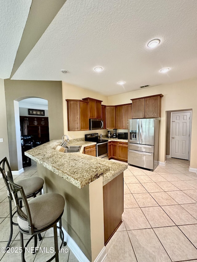 kitchen featuring light tile patterned flooring, a peninsula, a sink, appliances with stainless steel finishes, and a kitchen bar