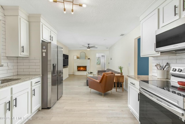 kitchen with stainless steel appliances, light wood-style flooring, a ceiling fan, a brick fireplace, and white cabinets