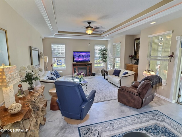 living room with ceiling fan, a tray ceiling, a glass covered fireplace, and baseboards