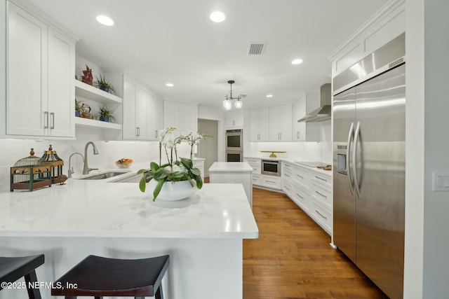 kitchen featuring a sink, open shelves, stainless steel appliances, a peninsula, and wall chimney range hood