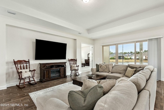 living room with a tray ceiling, wood finish floors, visible vents, a glass covered fireplace, and baseboards