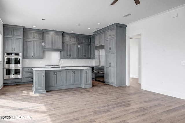 kitchen featuring a center island with sink, light wood finished floors, gray cabinets, light countertops, and appliances with stainless steel finishes