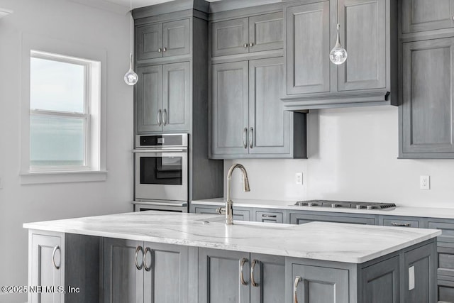 kitchen featuring a center island with sink, light stone counters, gray cabinets, stainless steel gas cooktop, and pendant lighting