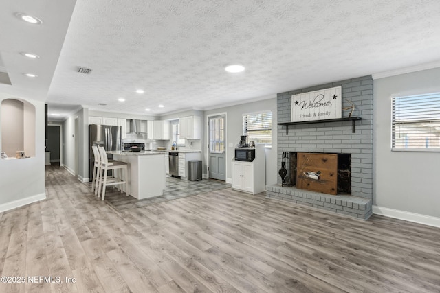 kitchen featuring light wood-style flooring, a textured ceiling, white cabinets, wall chimney range hood, and a brick fireplace