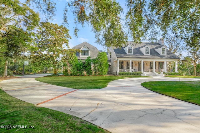 cape cod home featuring covered porch, concrete driveway, stone siding, a chimney, and a front yard