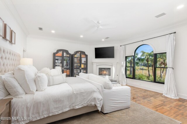 bedroom featuring a glass covered fireplace, visible vents, crown molding, and wood finished floors