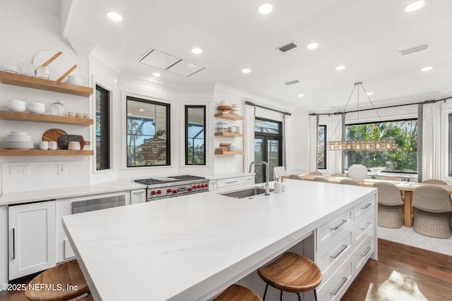 kitchen with open shelves, stove, white cabinetry, a sink, and an island with sink