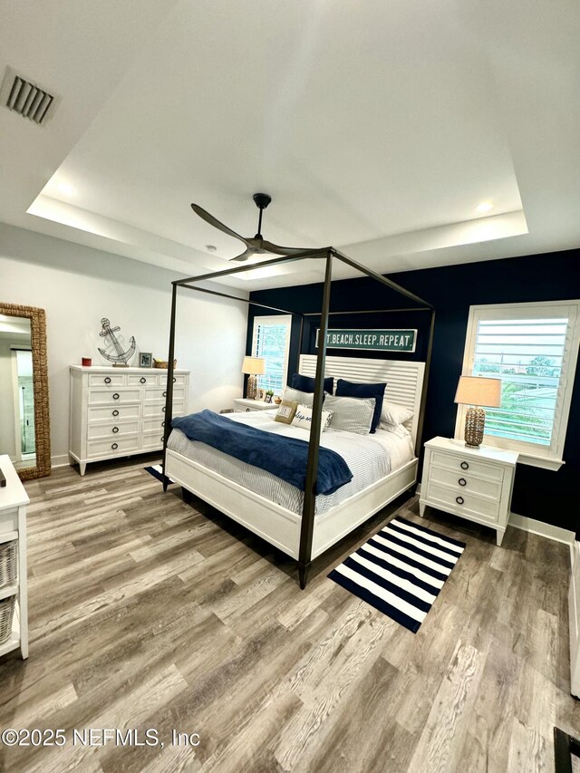 bedroom with a ceiling fan, wood finished floors, visible vents, baseboards, and a tray ceiling