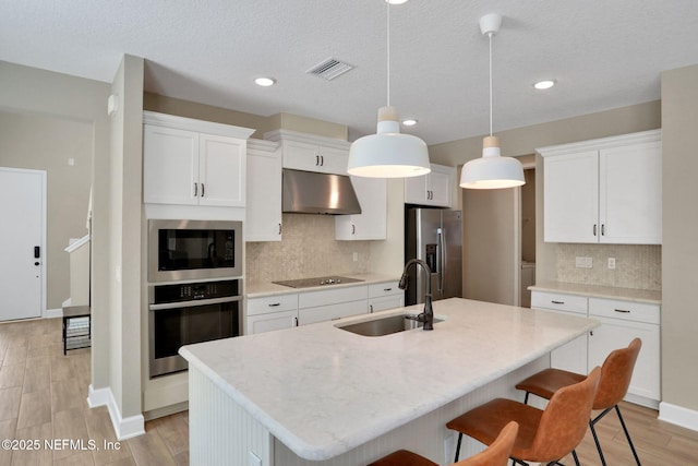 kitchen with a center island with sink, appliances with stainless steel finishes, a sink, under cabinet range hood, and a kitchen bar