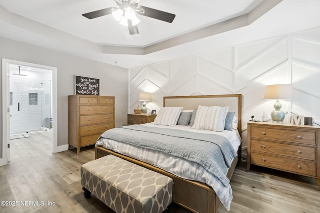 bedroom featuring a raised ceiling, a decorative wall, light wood-style floors, connected bathroom, and ceiling fan