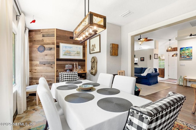 dining space featuring ceiling fan with notable chandelier, visible vents, wooden walls, and wood finished floors