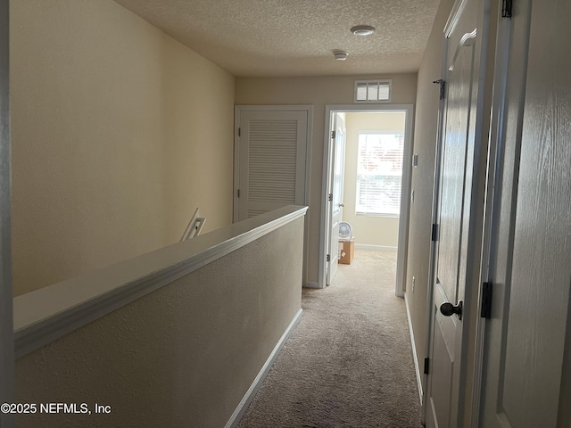 hall with a textured ceiling, light colored carpet, visible vents, baseboards, and an upstairs landing