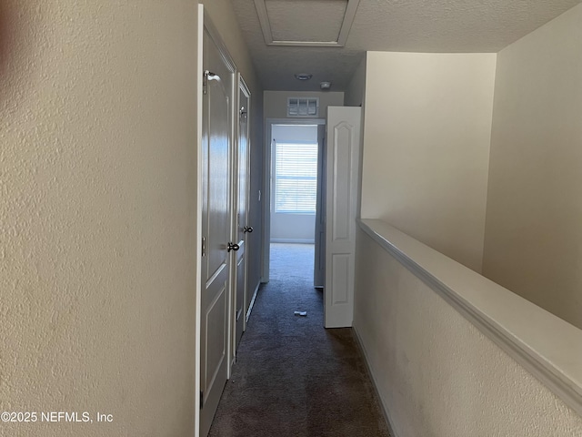 hallway with a textured ceiling, a textured wall, dark carpet, and visible vents