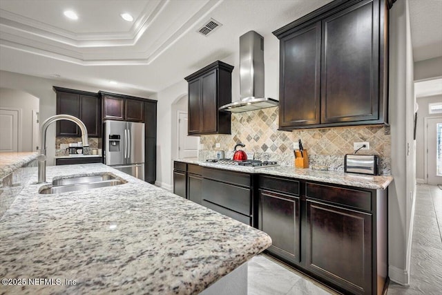 kitchen with arched walkways, a sink, appliances with stainless steel finishes, a raised ceiling, and wall chimney range hood