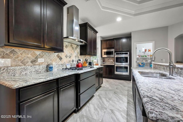 kitchen featuring tasteful backsplash, a tray ceiling, stainless steel appliances, wall chimney exhaust hood, and a sink