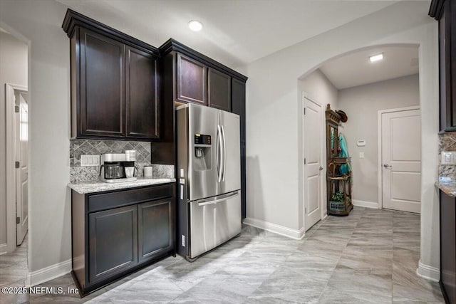 kitchen with stainless steel refrigerator with ice dispenser, backsplash, arched walkways, baseboards, and dark brown cabinets