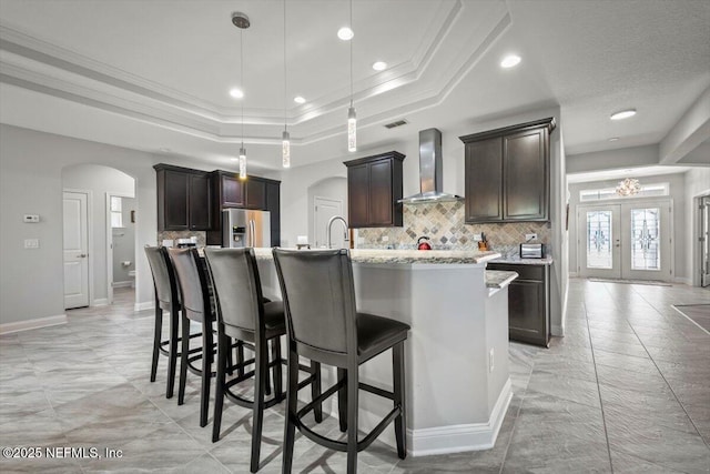 kitchen with a raised ceiling, wall chimney range hood, arched walkways, and stainless steel fridge