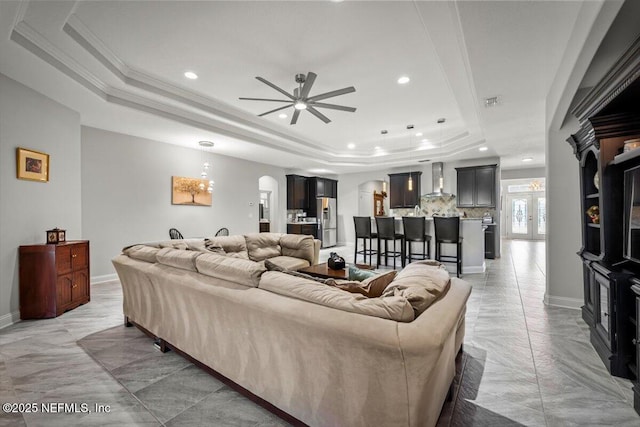 living area featuring a raised ceiling, ornamental molding, a ceiling fan, arched walkways, and baseboards