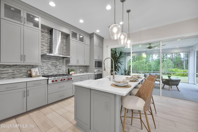 kitchen featuring stainless steel appliances, backsplash, gray cabinetry, wall chimney range hood, and an island with sink